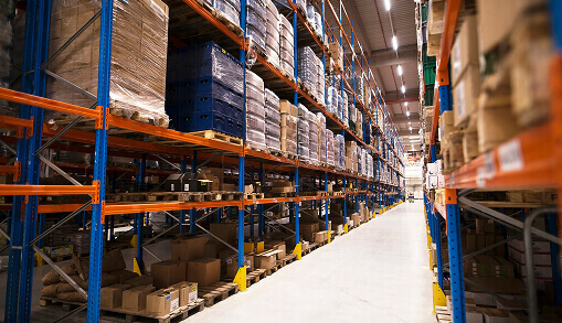 Pasillo de una bodega con estanterías azules y naranjas llenas de cajas y estibas hasta el techo
